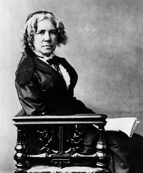 Black-and-white studio portrait of a seated woman in a dark dress, curled hair, papers at her side on a carved chair.