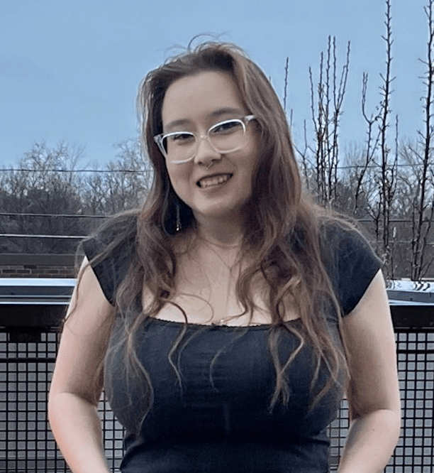 A photo of Misha Wichita, wearing glasses and a dark top, smiling while standing on an outdoor balcony with bare trees and a muted sky in the background.