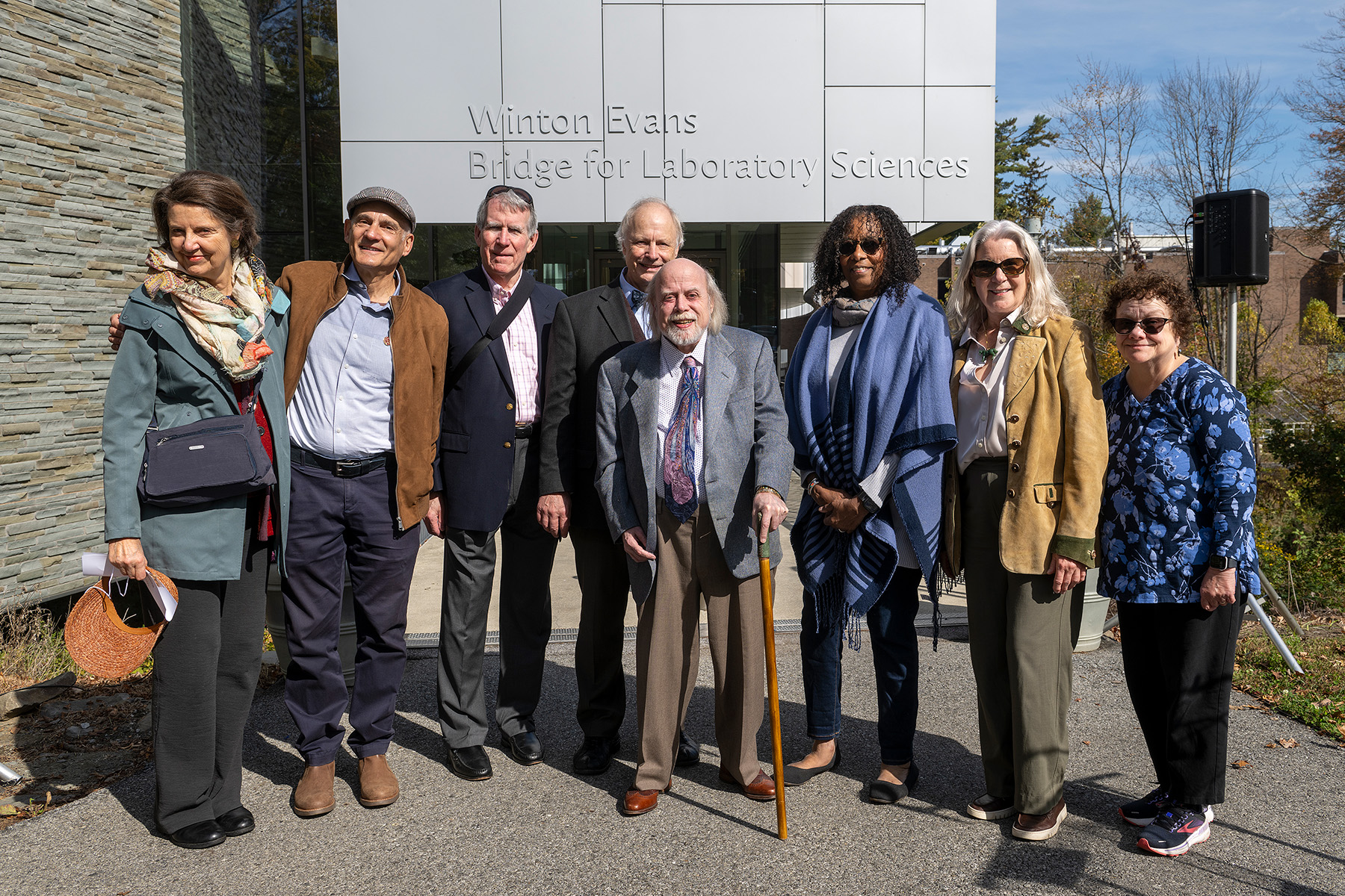 Eight adults stand together, smiling, in front of a campus building sign that reads “Winton Evans Bridge for Laboratory Sciences”; the person in the center holds a cane, and fall trees are visible in the background.
