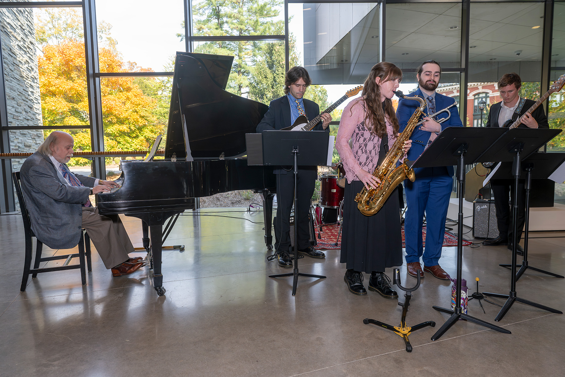Pianist seated at a grand piano performing with a small jazz combo—saxophonist, guitarist, bassist, and drummer—inside a bright glass-walled atrium, with music stands in front and autumn trees visible outside.