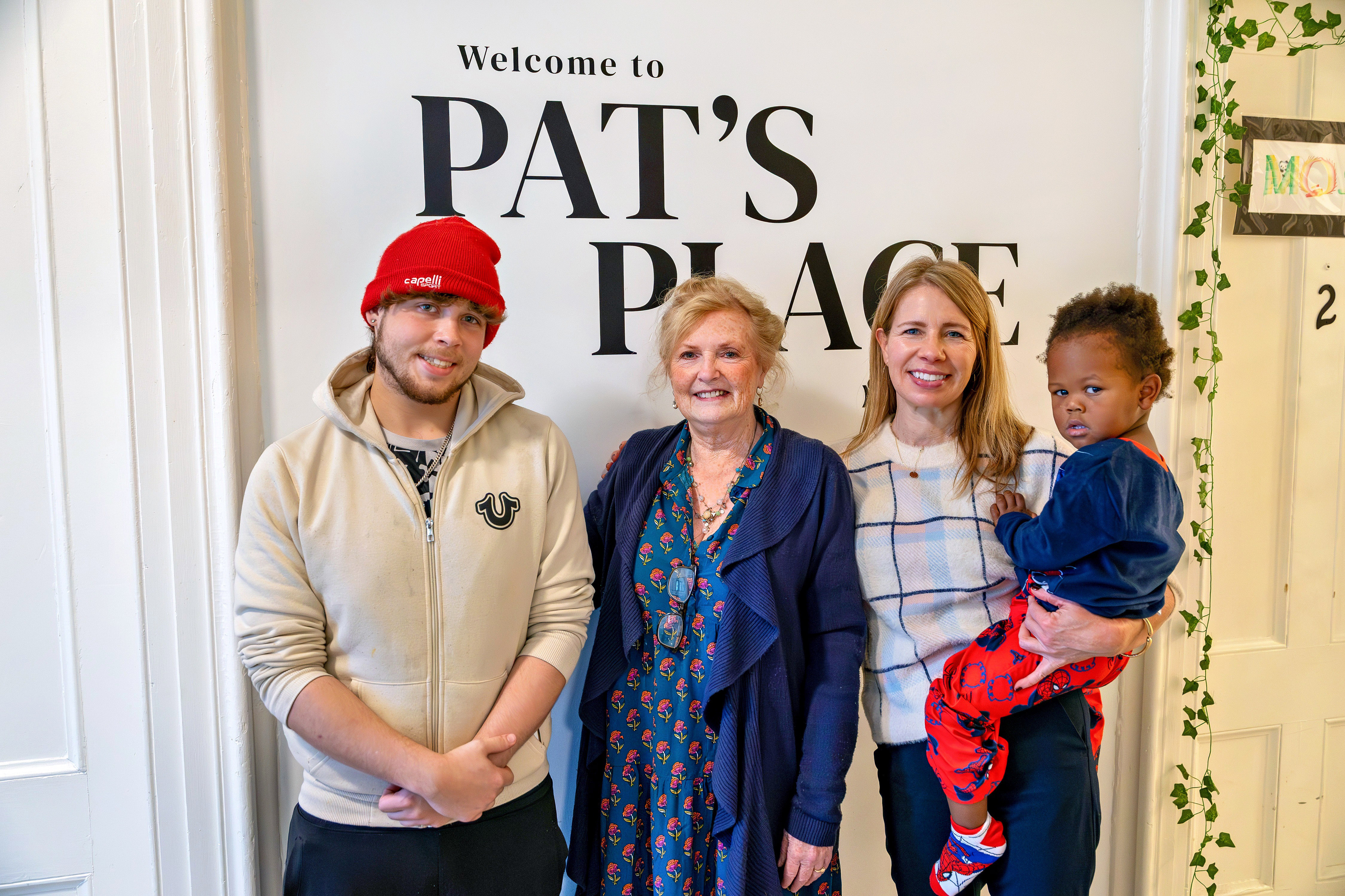 Three adults and one child pose indoors in front of a wall sign reading ‘Welcome to Pat’s Place’.