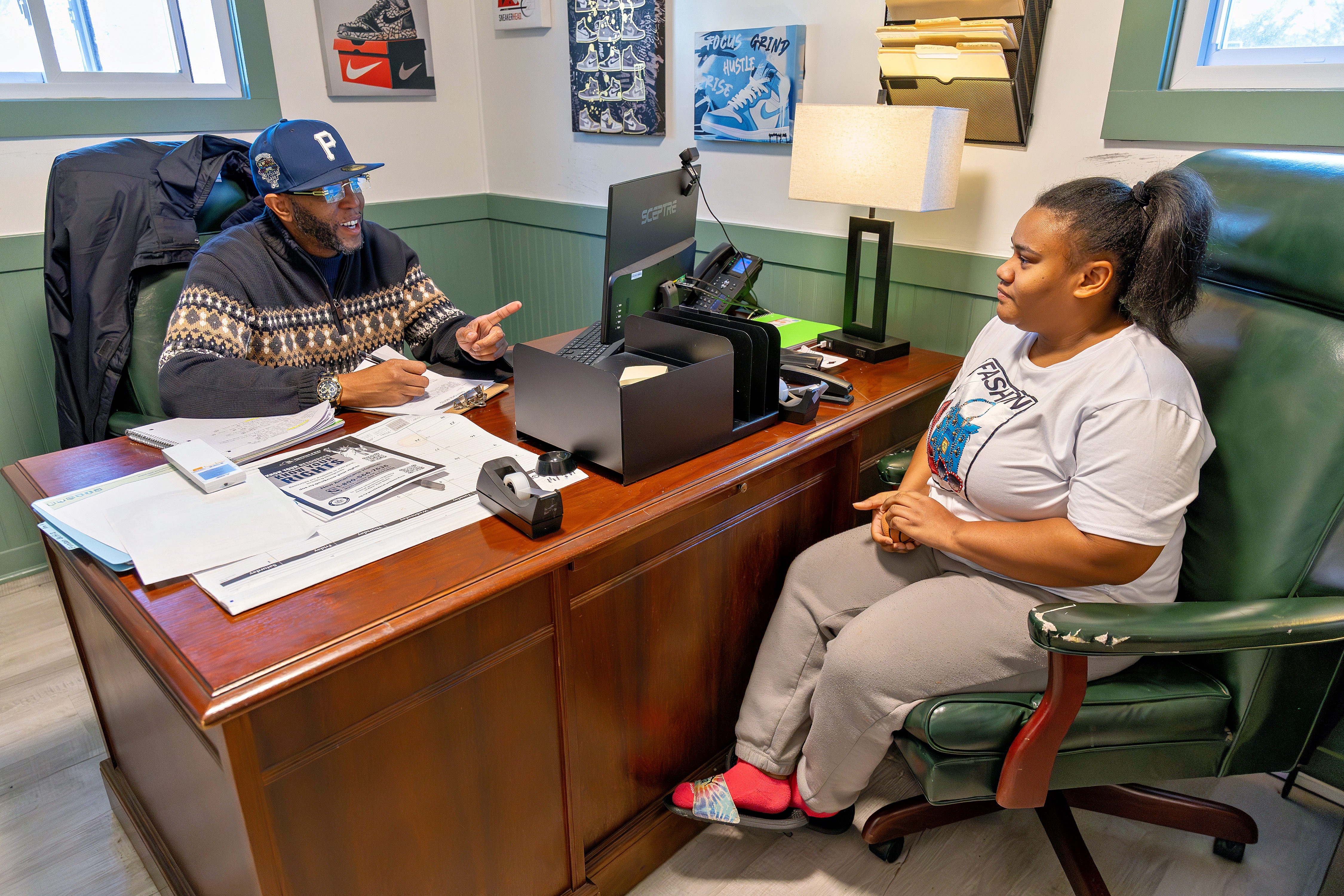 Two people sit across a desk in an office, talking; one person gestures while holding paperwork, and the other listens from a green chair.