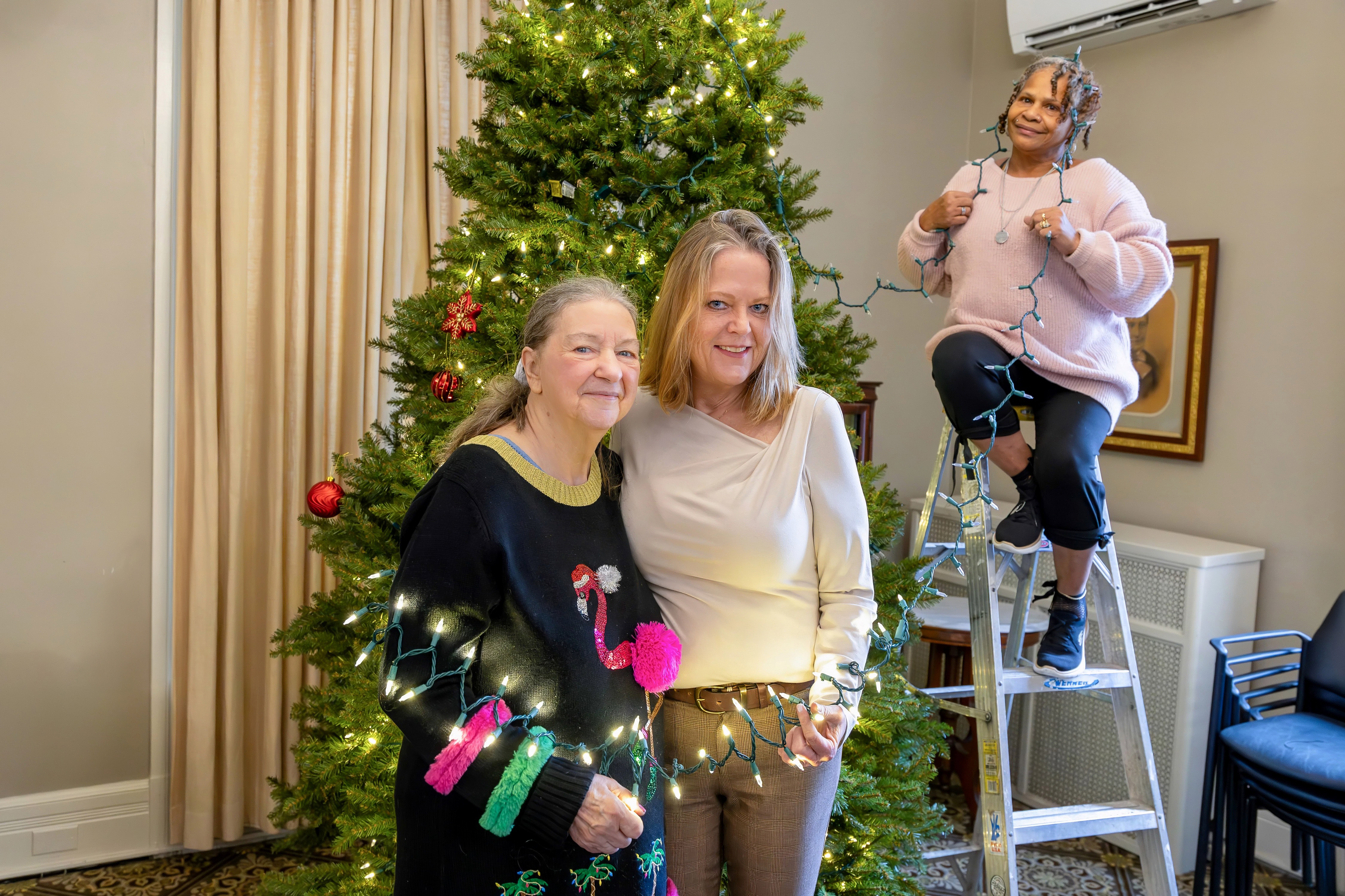 Three people decorate a large Christmas tree indoors; two stand smiling in front while a third stands on a step ladder holding a string of lights.