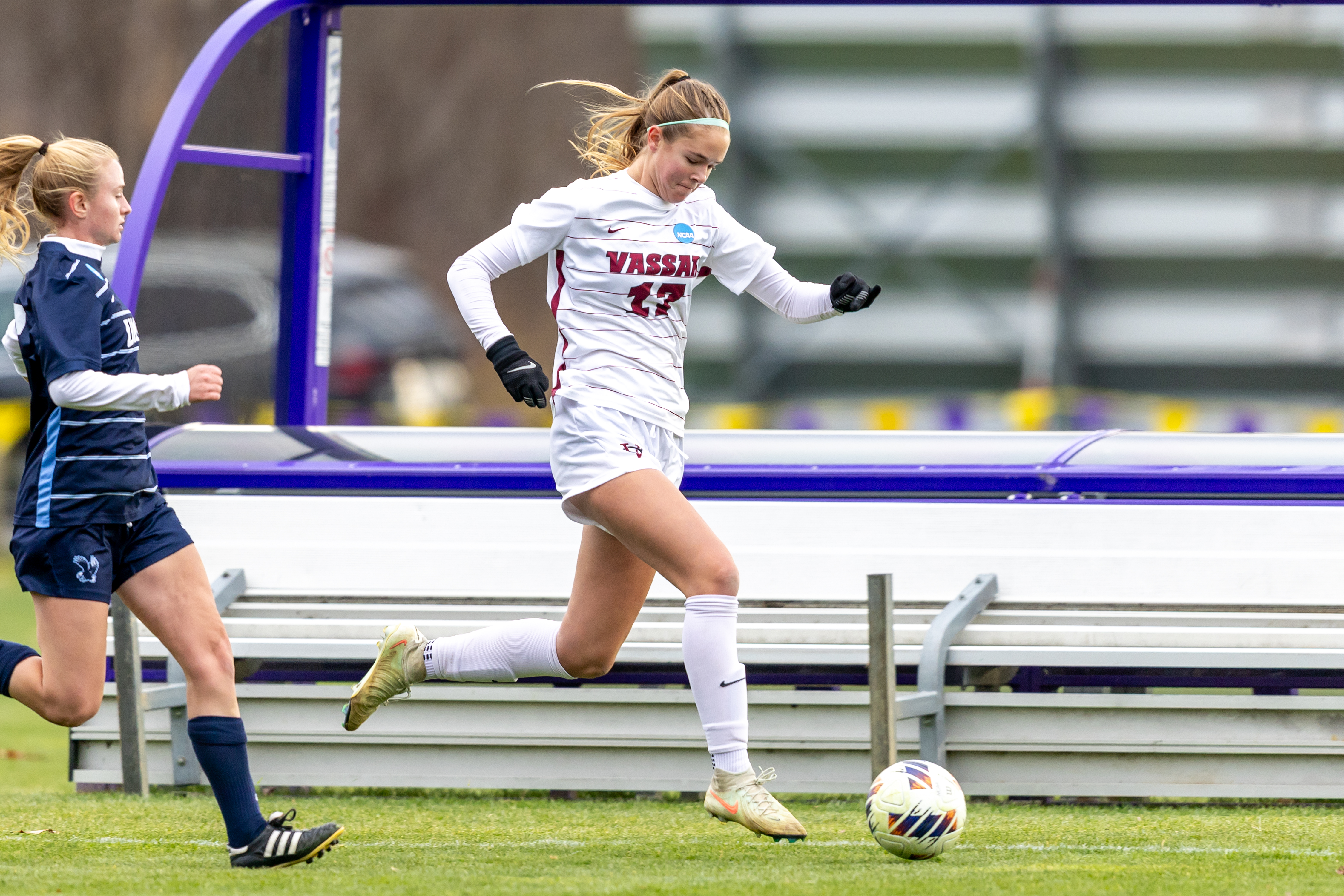 A soccer player wearing a Vassar jersey charges toward the open ball as an opposing player closes in from behind.
