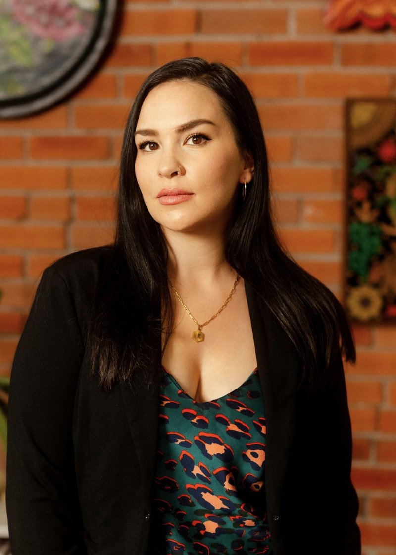 Kali Fajardo-Anstine standing in front of a short bookcase and a decorated brick wall.