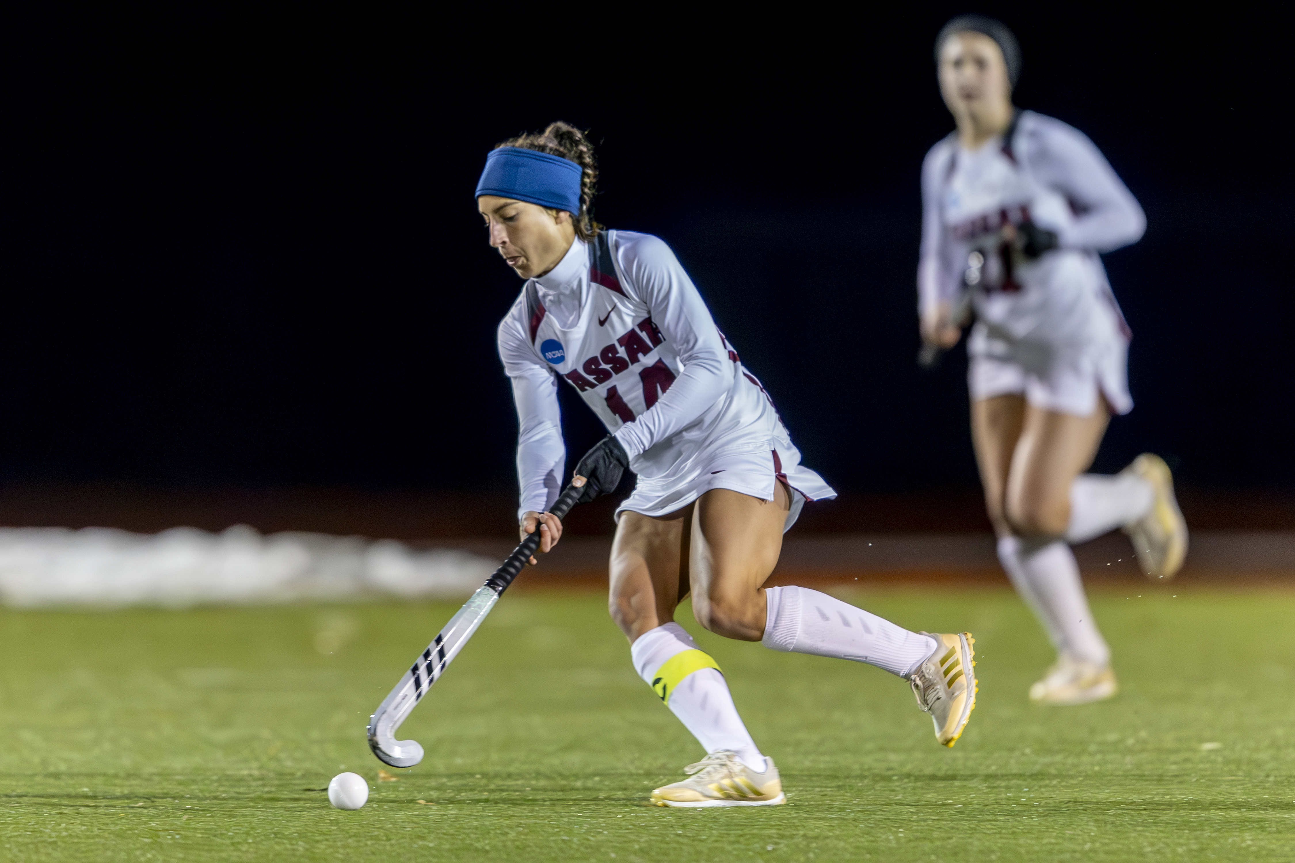 A field hockey player wearing a headband and Vassar jersey maneuvers the ball down the field with her stick.