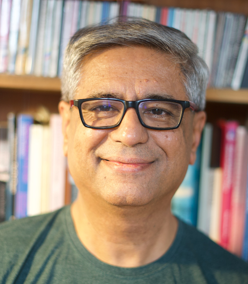 Smiling person with short gray hair and black glasses, wearing a green T-shirt, in front of a bookshelf with colorful books.