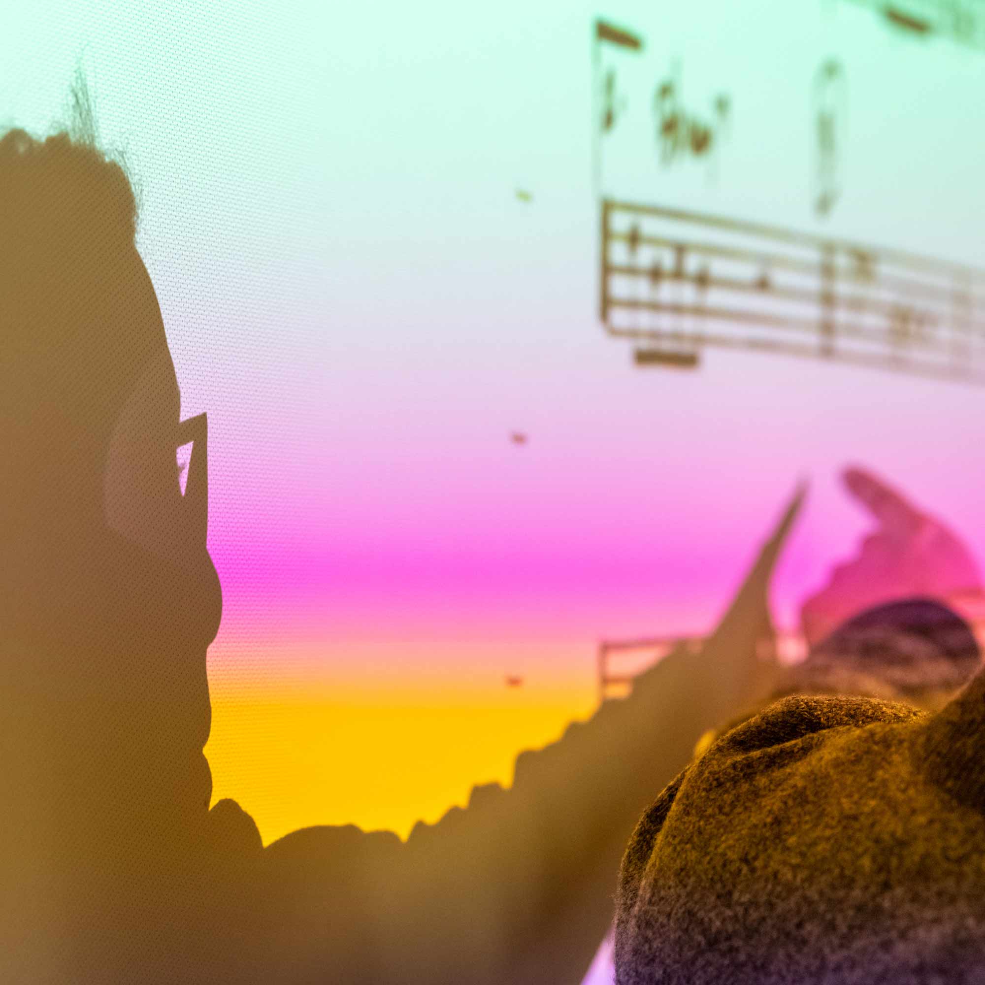  A person's silhouette is visible in the foreground, facing right, wearing glasses, and pointing toward music notation on a large screen or monitor. The screen displays a vivid, pixelated gradient background of yellow, pink, and bright cyan.