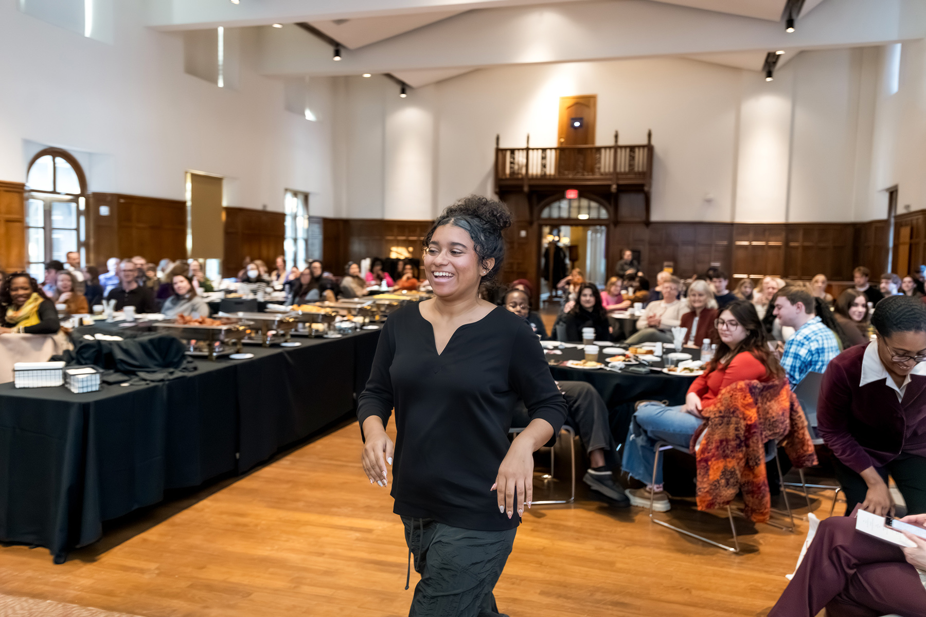 A smiling attendee walks through a large hall as seated guests watch, with tables of food and a wood-paneled room in the background.