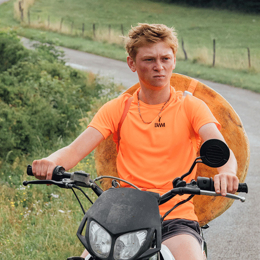 Person in a bright orange shirt rides a scooter on a country road with a giant wheel of cheese strapped to their back.