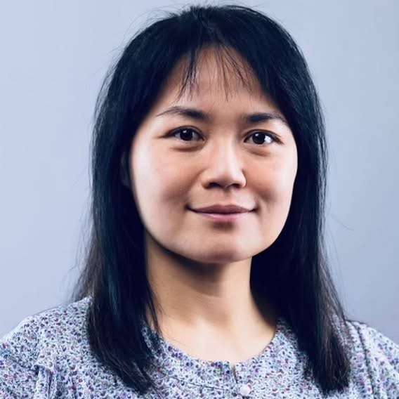 Headshot of a person with straight dark hair and bangs, wearing a patterned blouse, looking at the camera with a slight smile against a plain light gray-blue background.