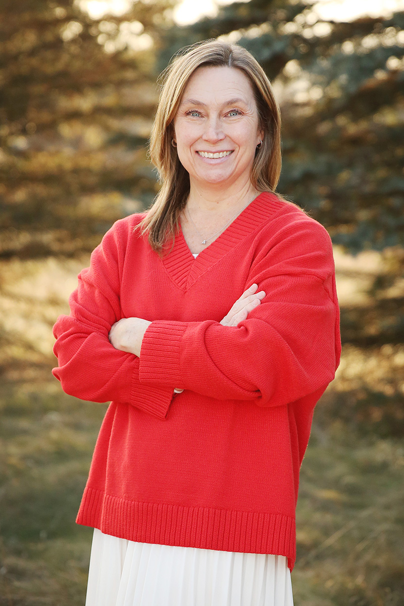 Amy Allen stands outdoors with her arms crossed, smiling, wearing a red sweater against a softly blurred natural background.