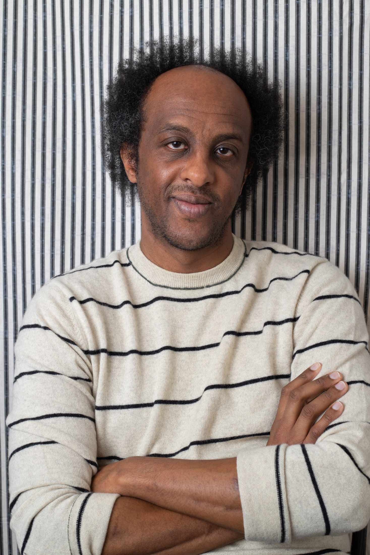 A person with dark curly hair and a slight smile poses with arms crossed. They wear a light-colored sweater with thin horizontal black stripes against a background of vertical black and white stripes.