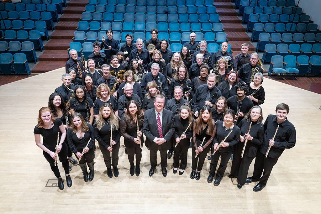 A group portrait of the wind ensemble members with instruments in concert dress on the stage.