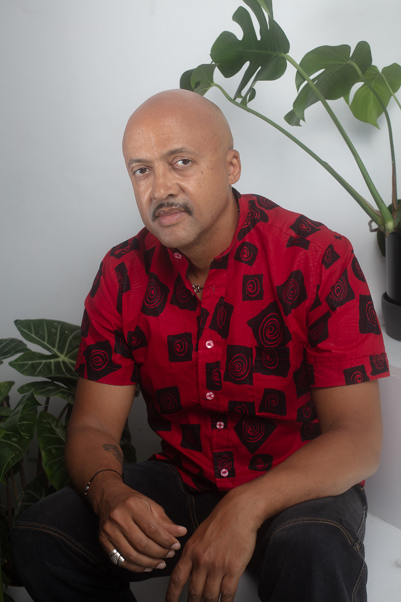A portrait of an individual wearing a red and black patterned shirt, sitting between two potted plants against a neutral white wall.