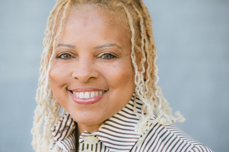 Smiling person with light blond locs wearing a striped collared shirt, against a soft blue background.