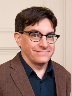 A photo of Jamie Kelly, a person with dark brown hair, glasses, and a brown suit coat.