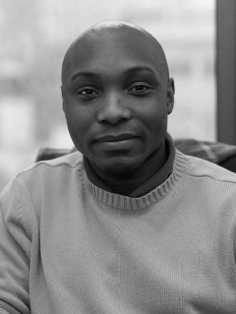 A portrait of Dr. Kafui Attoh, a person with a shaved head and a neutral expression, looking directly at the viewer. They wear a light-colored sweater with a ribbed collar, which adds texture to the image. The background features soft focus, creating an out-of-focus effect that enhances the clarity of the subject against a subtle, cloudy environment.