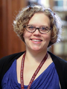 A photo of Kira Thompson. They have blond curly hair, glasses, and a blue top. A lanyard with "Poughkeepsie Public Library District" printed on it hangs around their neck.