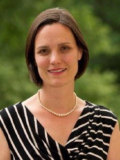 A portrait of Laura McCann, a person with shoulder-length brown hair, styled straight and parted slightly off-center. They wear a black and white striped dress and a pearl necklace.