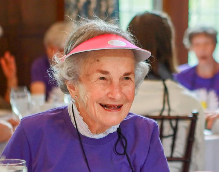 A person with short light gray hair smiling while seated at a table, wearing a red visor and a purple shirt at a Vassar Reunion luncheon.