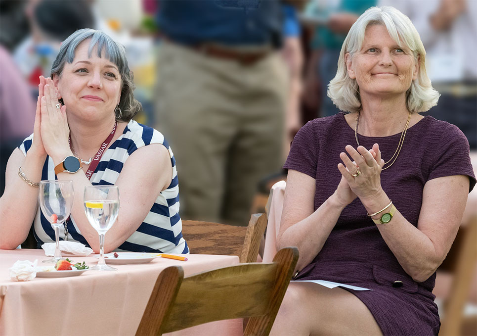 Vassar President Bradley sits at a table with another person, clapping.