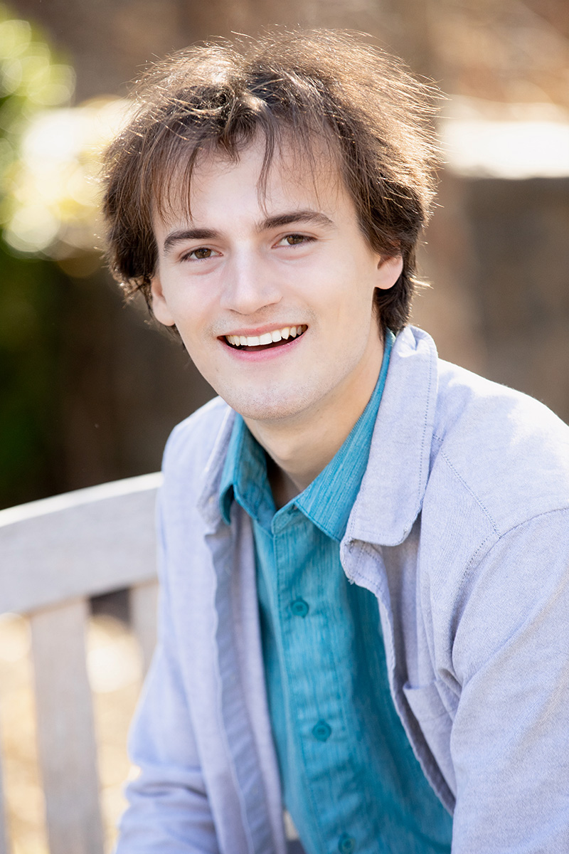 A person with brown hair smiling brightly while sitting outdoors on a wooden bench.