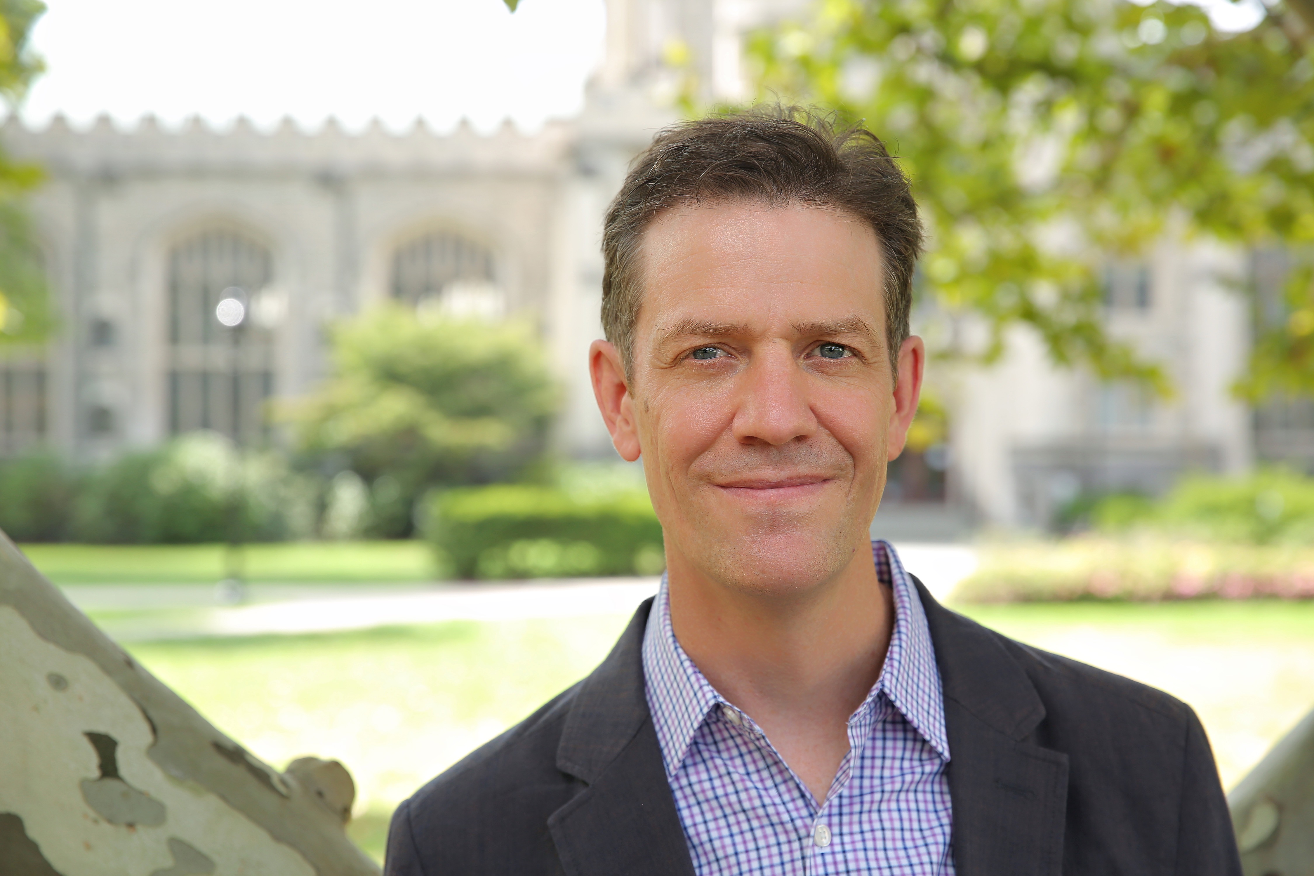 A smiling person wearing a collared shirt and blazer stands on the Vassar campus with the college library in the background.
