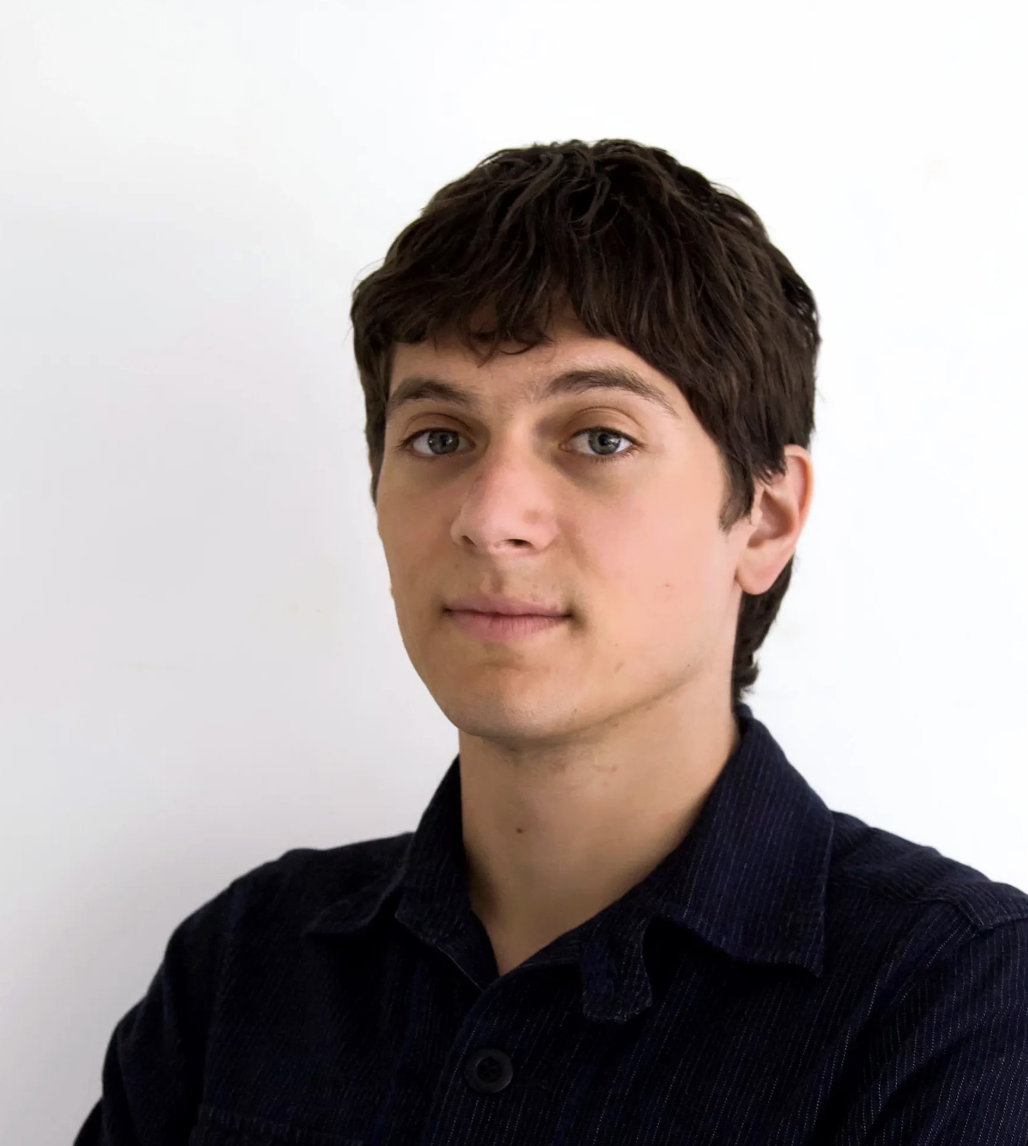 A close-up portrait features a figure standing against a plain white wall. The figure has short, dark hair and a neutral expression, with arms crossed in front. They are wearing a dark-colored button-up shirt, which adds texture against the smooth surface behind them. The lighting is soft and even, highlighting the features and creating a balanced appearance.