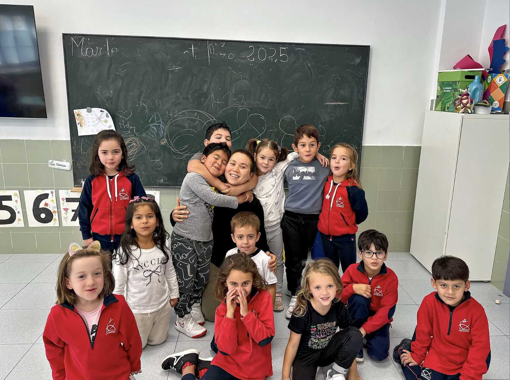 A group of children stands together in a classroom, arranged in two rows. The front row features four children sitting on the floor, while the back row includes several others standing. Several are hugging an adult with brown hair. The walls are adorned with educational materials, including a chalkboard with handwritten notes and numbers. The scene is brightly lit, with large windows contributing to the overall lightness of the room. The children are dressed in a mix of colorful clothes.