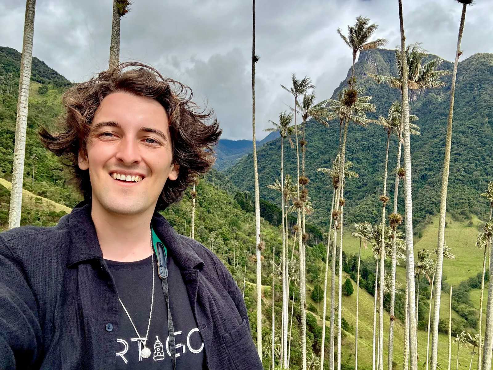 A person stands in a lush, mountainous landscape, surrounded by tall, slender trees. Bright green foliage contrasts with the cloudy sky, which features varying shades of gray. The person has brown hair, and they wear a dark, open jacket over a black shirt.