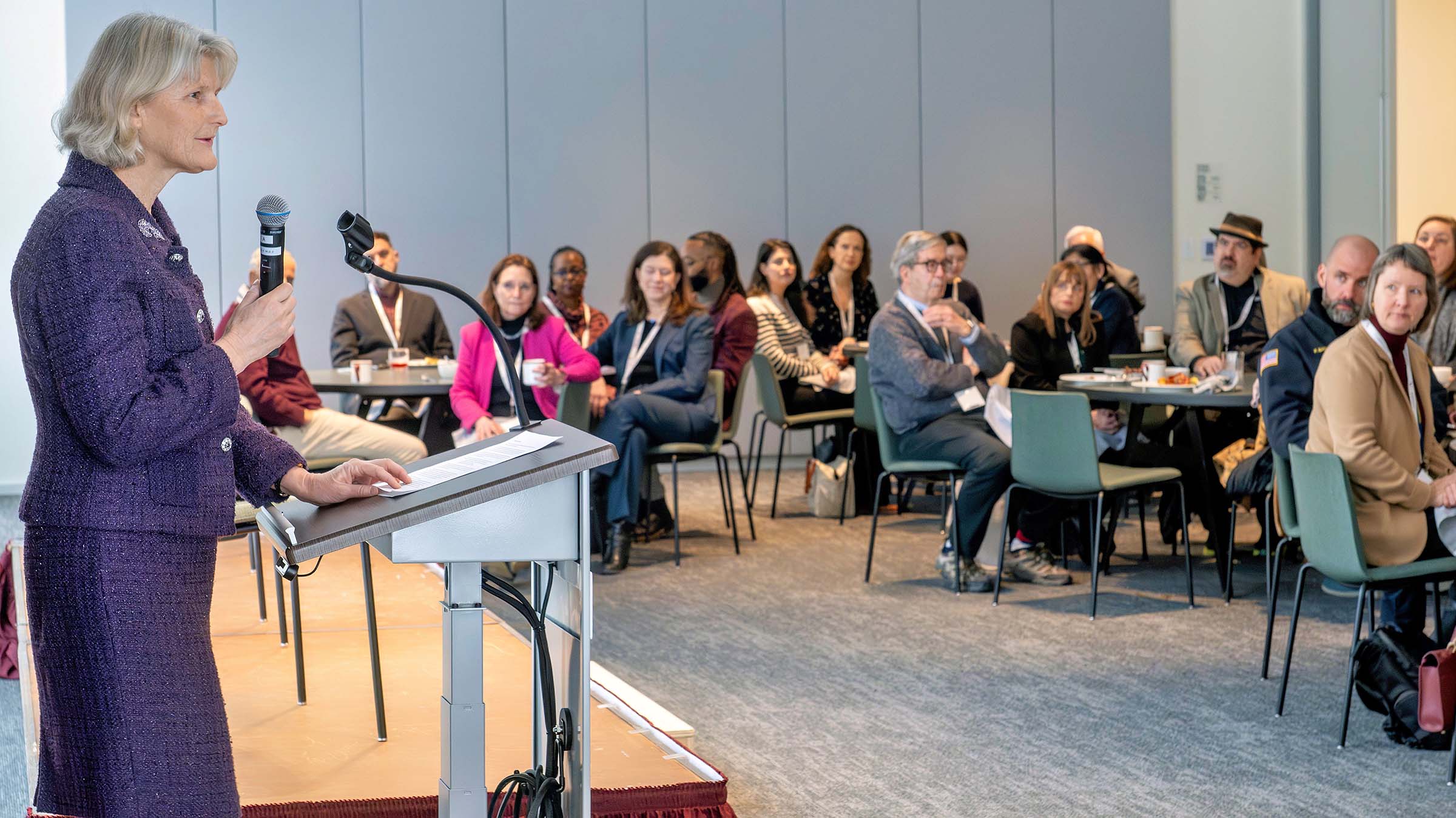 President Elizabeth H. Bradley in a purple suit stands at a podium speaking into a microphone to an audience seated at round tables at the the College’s inaugural Community Breakfast.