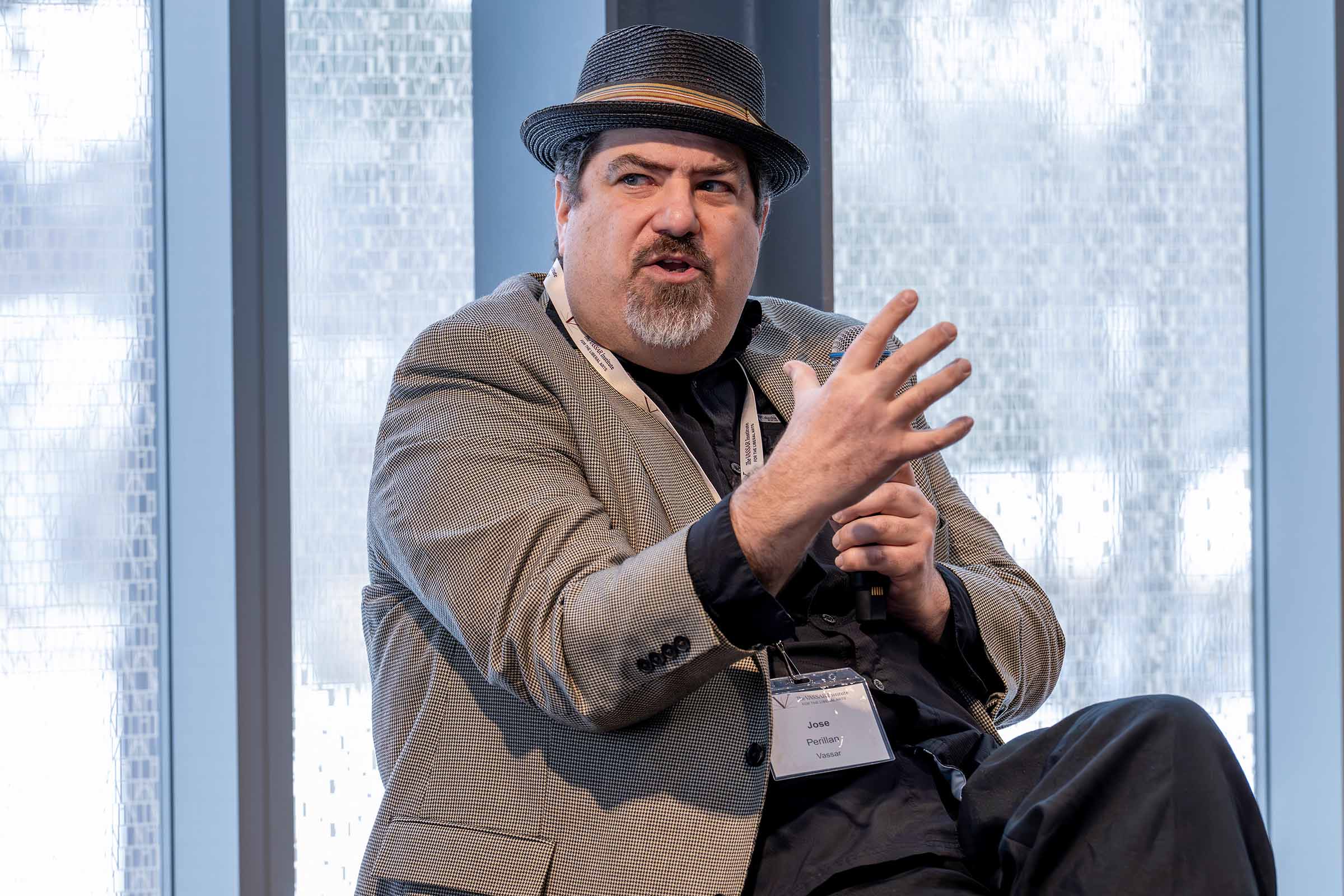 A man with a goatee and dark hair, wearing a grey patterned blazer, a dark shirt, dark pants and a dark fedora with a striped band. He is seated and gesturing with his right hand, while wearing a lanyard with a name tag that reads "Jose Perillan" from "Vassar".
