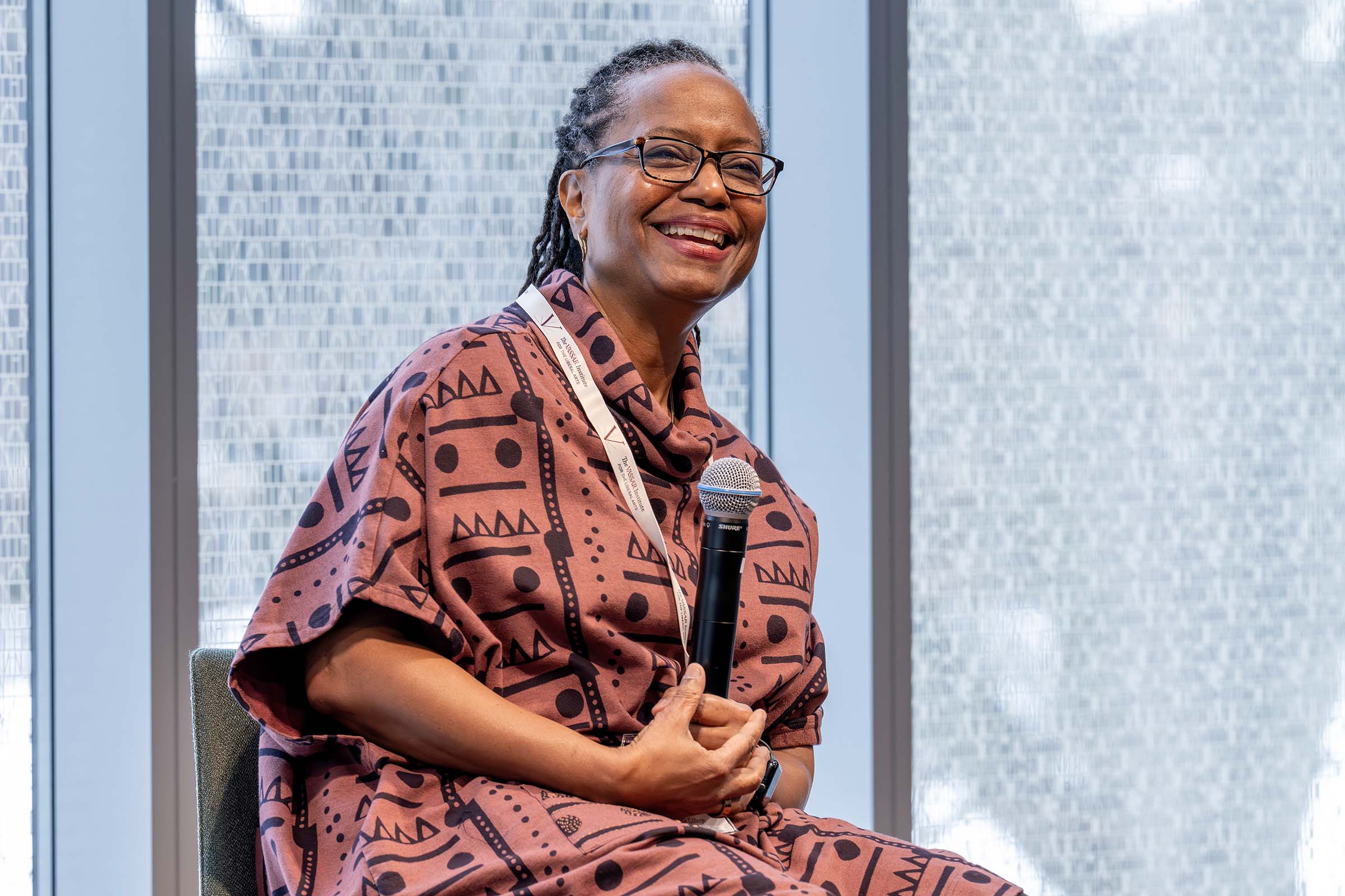 Candice M. Lowe Swift, with dark, salt-and-pepper braids and glasses is smiling broadly while seated. She is holding a black microphone and wearing a mauve-colored top with bold black geometric patterns and a white lanyard with a large, textured glass window in the background.