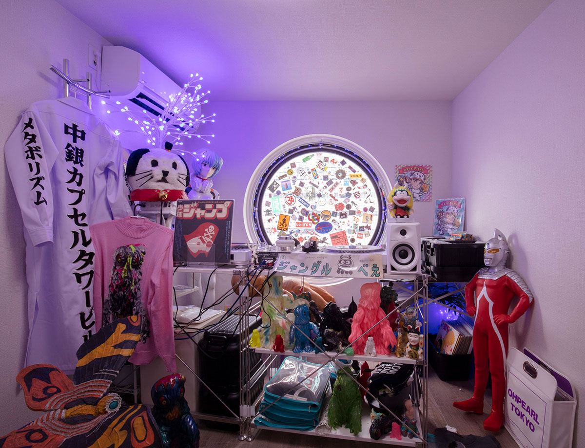 A compact, capsule-like room filled with colorful Japanese pop culture objects and media equipment, including a circular window covered with stickers, shelves of vinyl records and figurines, speakers and turntables, hanging garments with Japanese text, and a red Ultraman figure standing on the floor, illuminated by purple accent lighting.