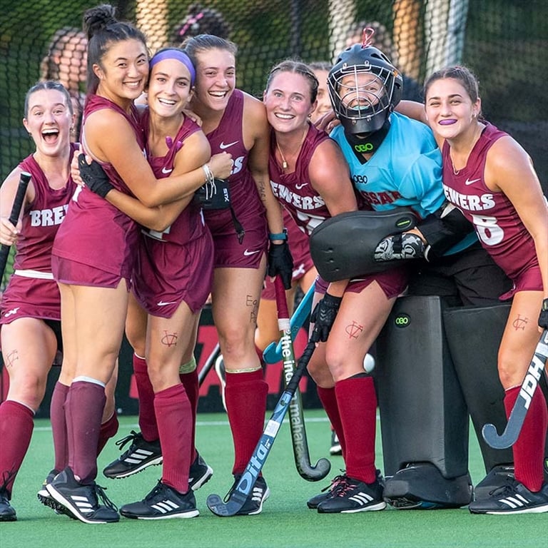 Vassar College field hockey players and their goalie smile and embrace on the field during a celebration.