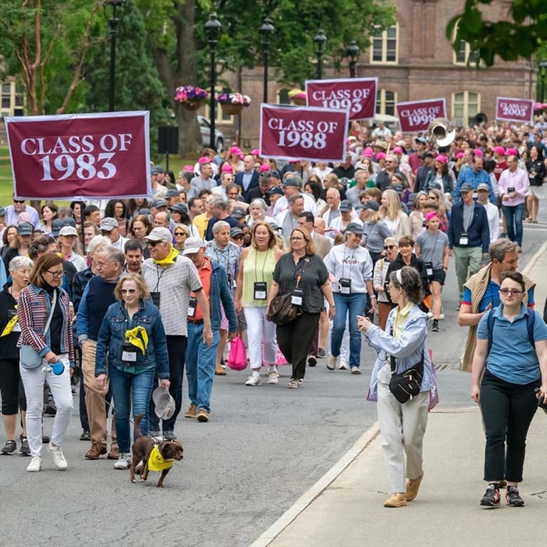A parade of many people wearing casual clothing walk along a road on Vassar's campus behind large red banners with various class years written on them.
