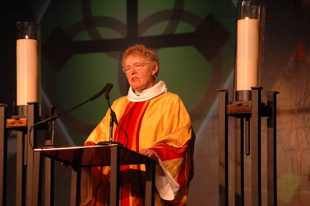 A person wearing eclesiatic attire stands at a pulpit framed by two large candles on stands.