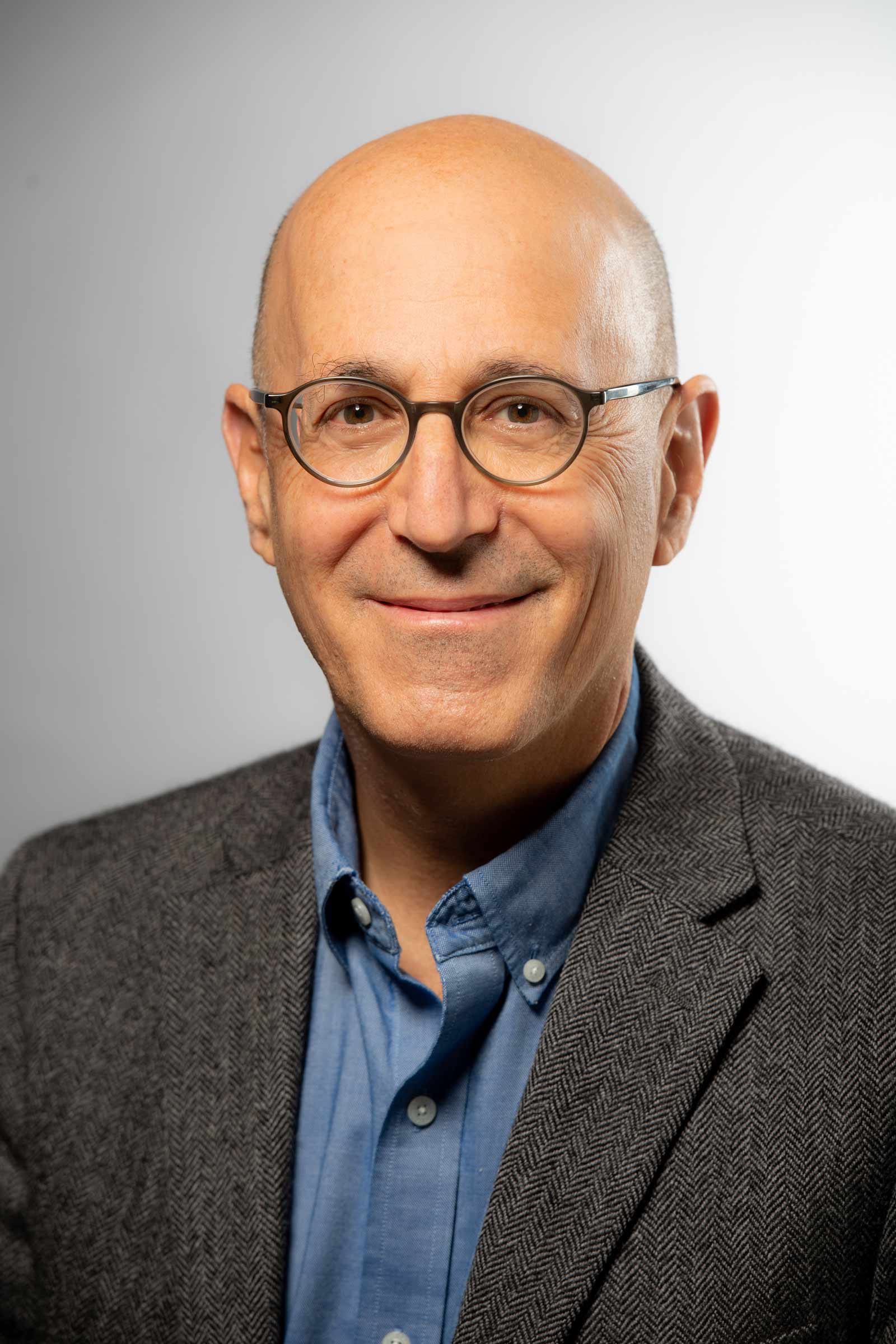 A bald individual with round glasses and a slight smile faces forward. They wear a light blue button-down shirt underneath a grey herringbone textured blazer. The background is a solid, neutral light grey.