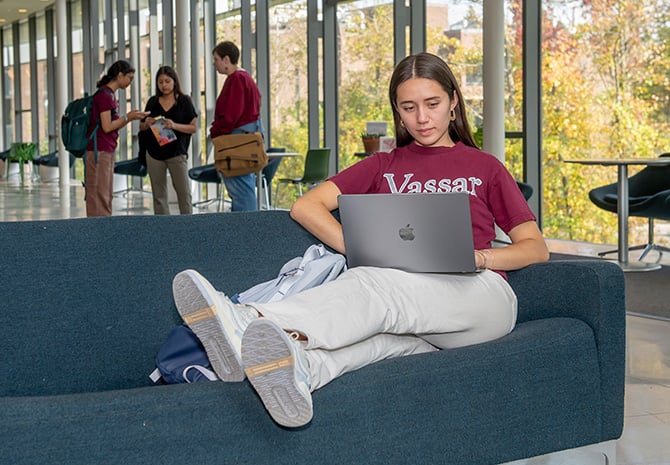 A student sits on a couch with a laptop open on their lap in a wide, windowed, curved hallway, with a group of students talking in a group in the background.