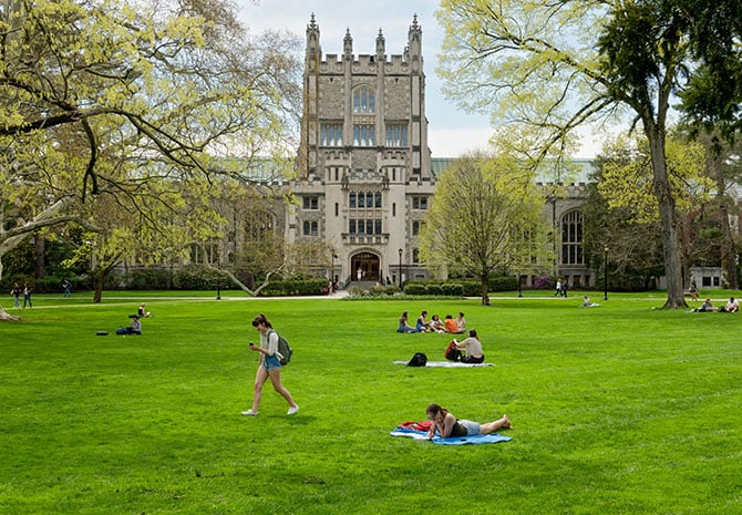 A gothic style building with a central tower and two wings on either side at the back of a large green lawn with students sitting or walking across it, with trees beginning to bud all around.
