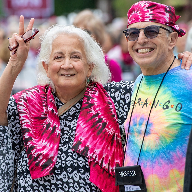 Two people wearing tie-dyed shirts, one flashing the peace sign with their right hand while holding a set of glasses, smiler at the viewer.