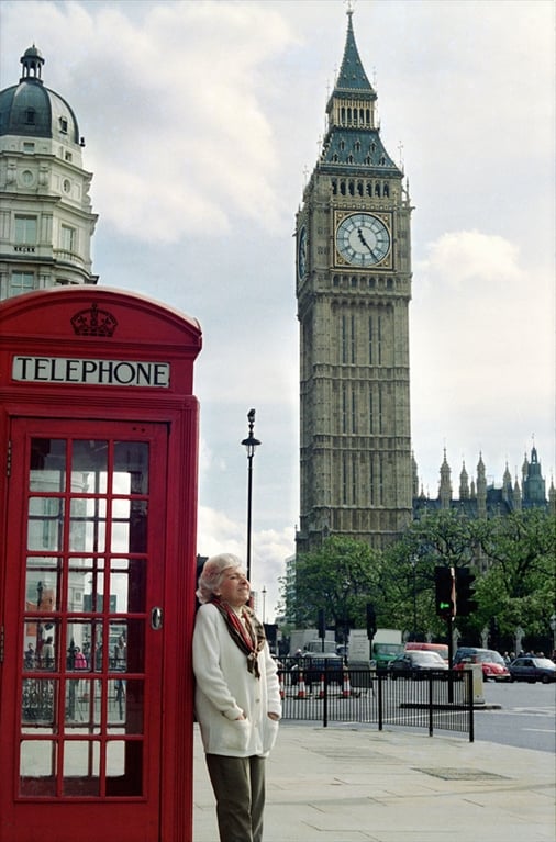 A person with short white hair wearing a white sweater, colorful scarf, and dark colored pants leans against a classic red telephone booth in London, with Big Ben and the Houses of Parliament in the background.