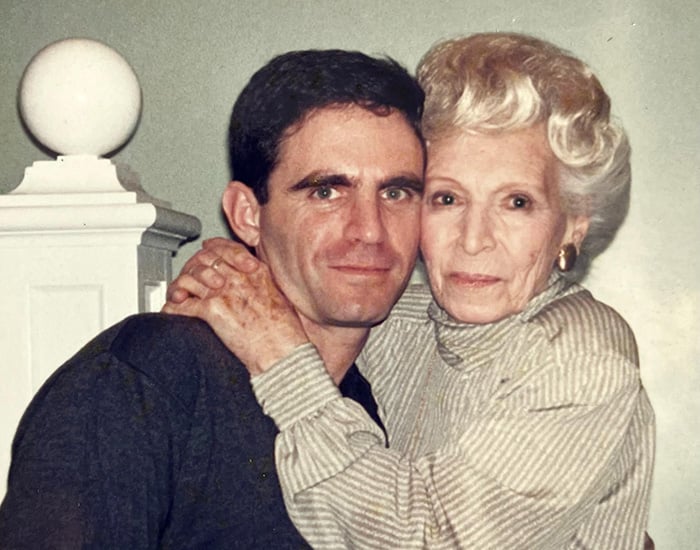 Two people embrace closely in a warmly lit interior space. One person with short silver/blond wearing a beige shirt has their arm around the other per
