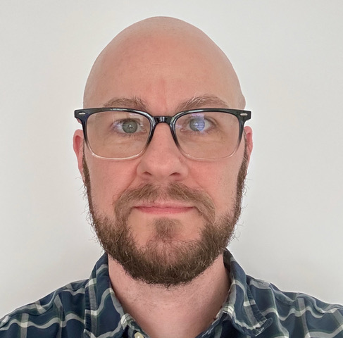 A bald individual with a brown beard and mustache wears black-rimmed glasses. They are wearing a blue, green, and white plaid button-down shirt and looking directly at the camera against a plain, off-white background.
