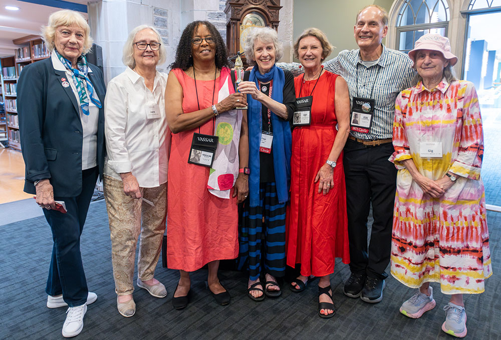 A group of seven people stand together inside a library wearing name tags and lanyards. One person in the center is holding a small trophy.