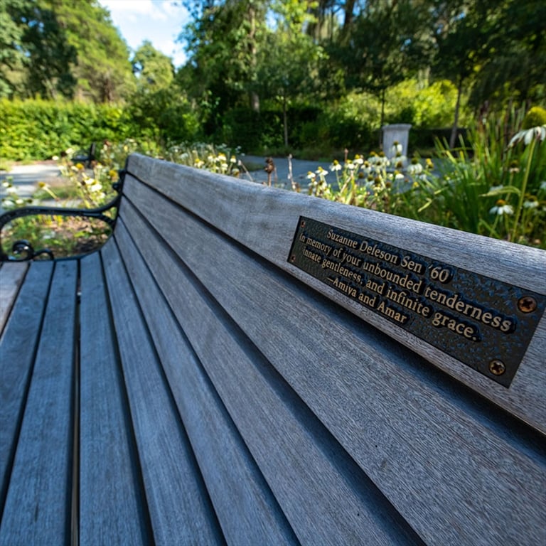 A view of a plaque on the back of a wooden bench in a garden-like setting, which has a person's name, class year, and a memorial quote.