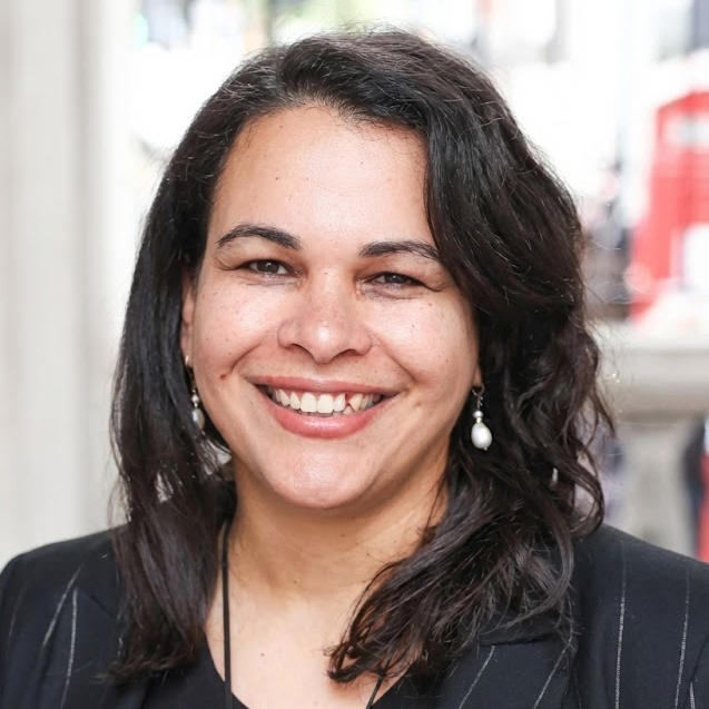 Smiling individual with dark wavy hair wearing a black top and a dark pinstriped blazer. They are accessorized with pearl drop earrings and a large black and white geometric pendant necklace. The background is a brightly lit outdoor setting.