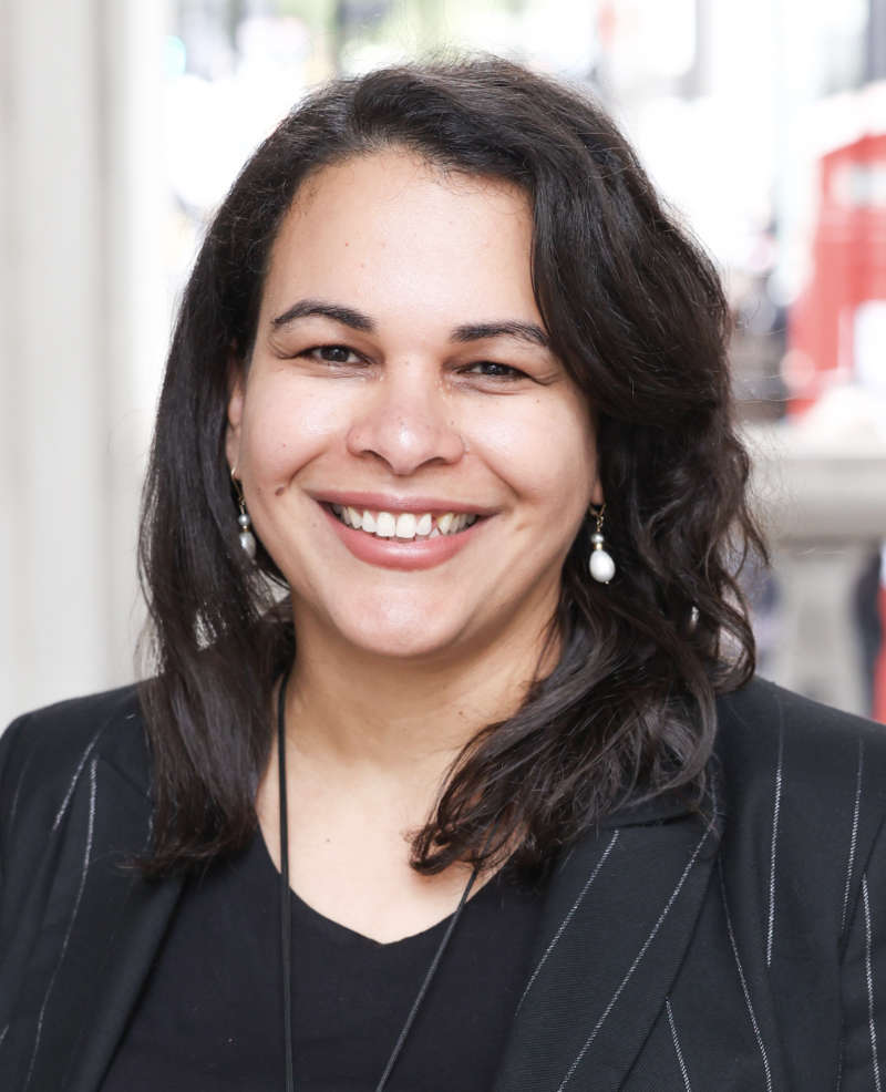 A photo of Rosa Andújar. They are smiling and wearing a dark blazer with subtle pinstripes and a black top.