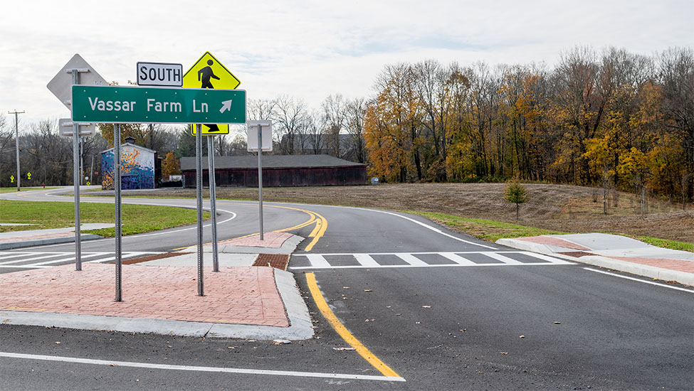 A newly constructed roadway with a pedestrian crossing and directional sign that reads Vassar Farm Lane, with a barn and autumn trees in the background